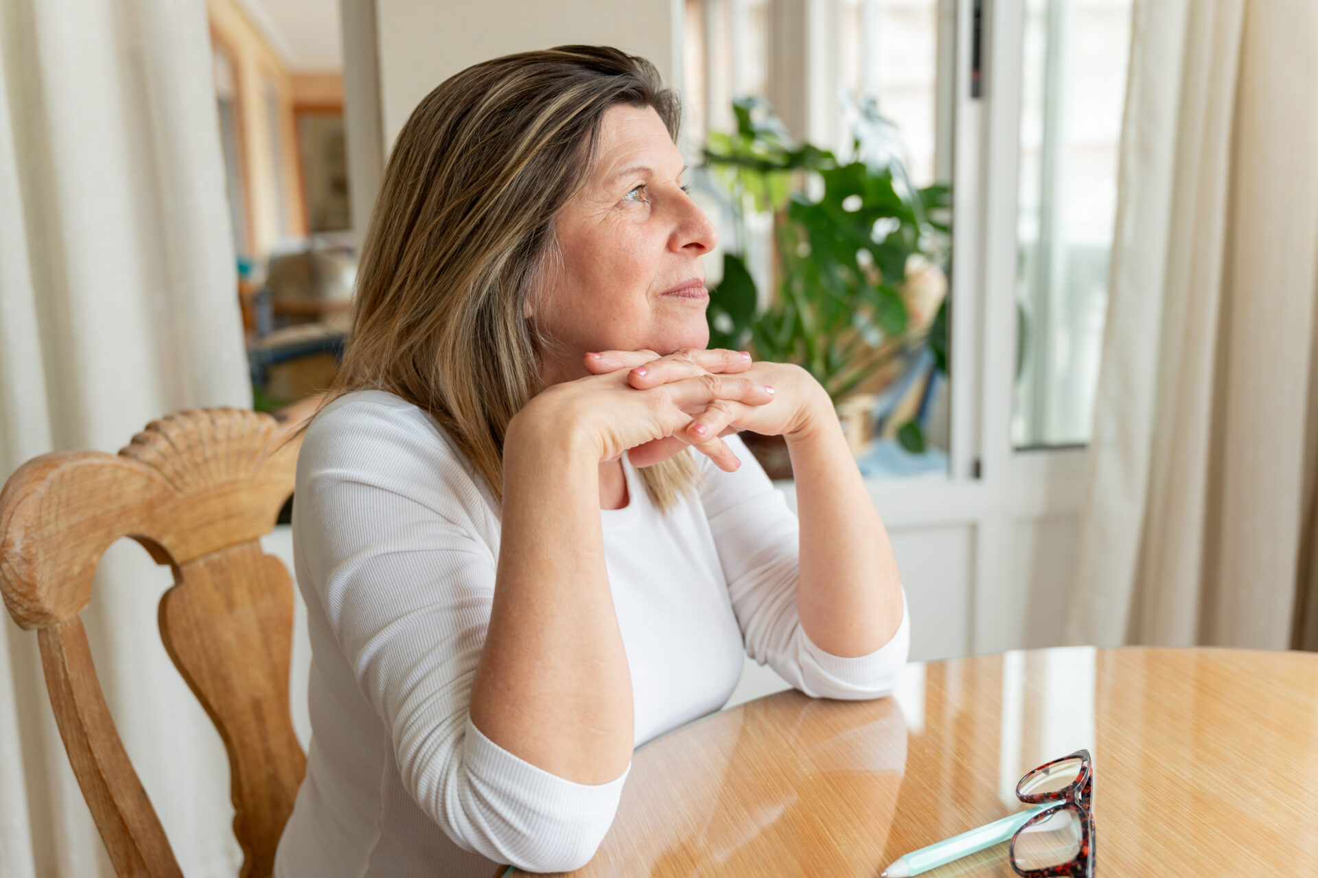 Mature woman resting hands clasped, pensively staring through window with reflective expression at wooden table